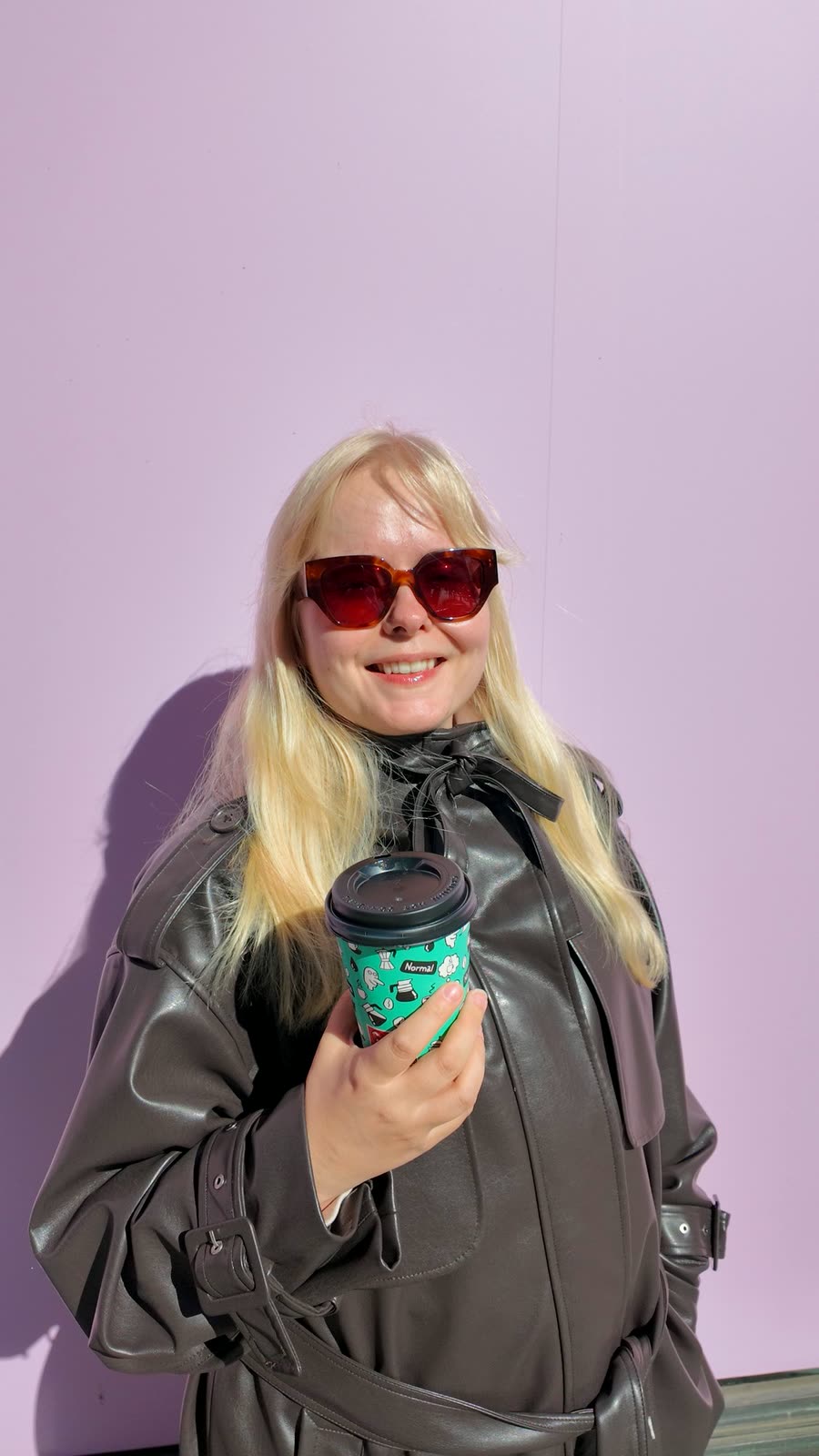 Aino smiling with a takeaway coffee in front of a lilac wall